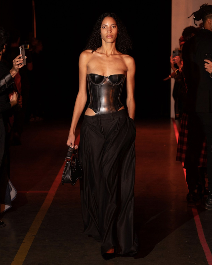 Slender female model with tan skin wearing a latex corset and black pants walking on a runway against a dark backdrop